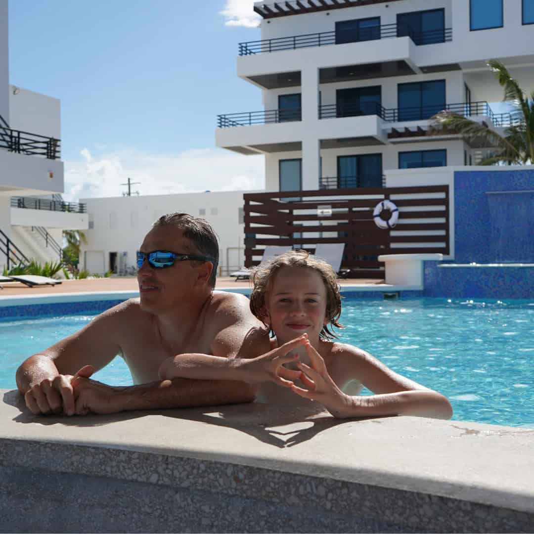 A man wearing sunglasses and a blonde boy with curly hair at the edge of a pool with a white hotel behind them.