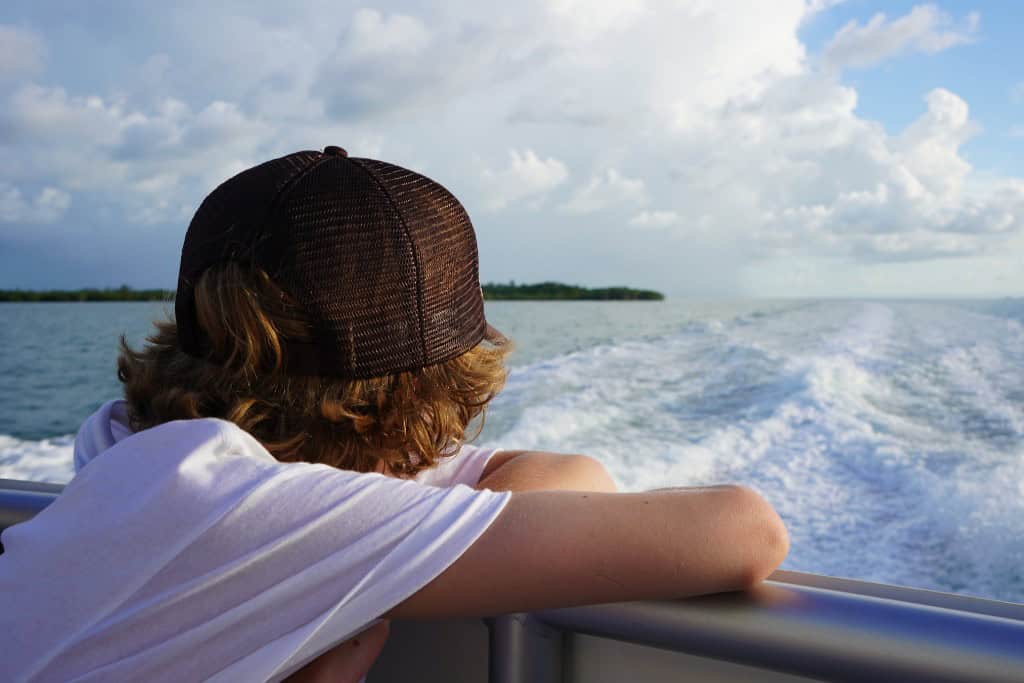 A boy in a brown baseball cap and white tee shirt looking out the back of a boat cruising through water with a tree colored island in the background.