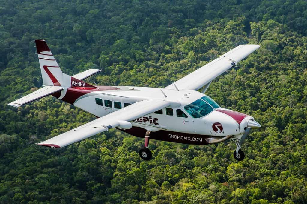 A small white plane with maroon markings that says Tropic Air on it flying above a tropical jungle.