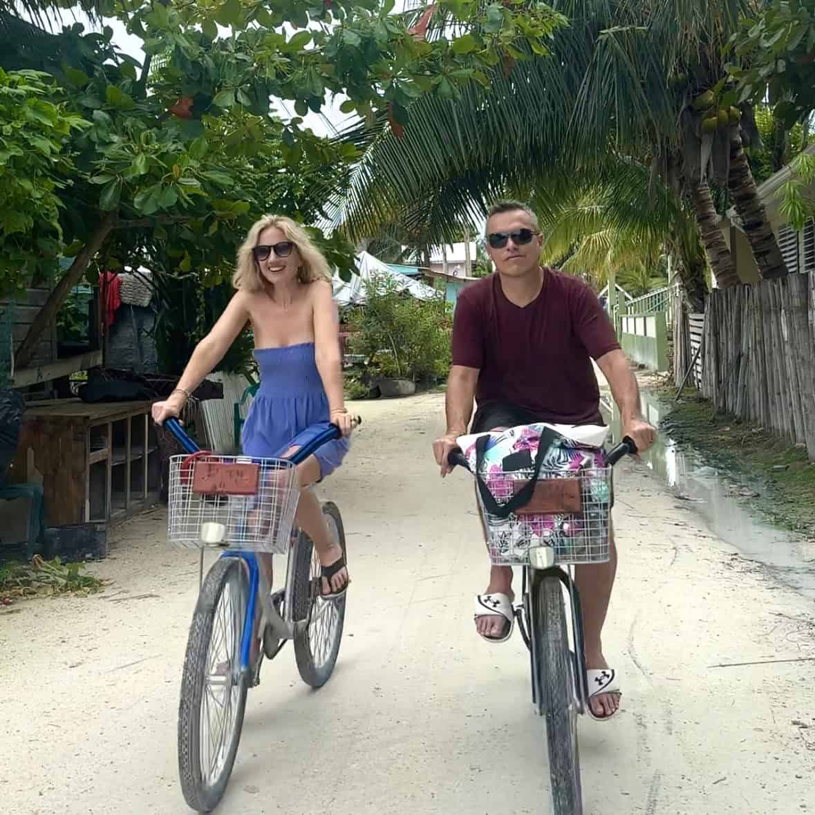 A blonde woman in a blue dress and sunglasses and man in a red shirt and sunglasses smiling and biking towards the camera.