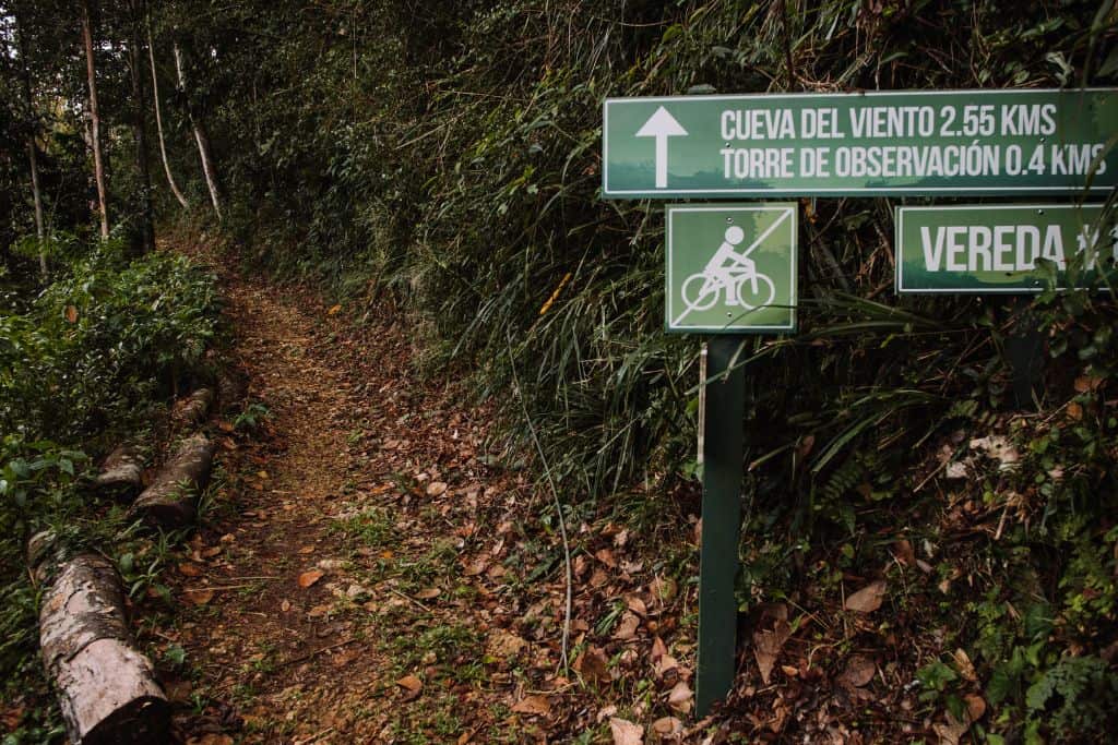 A dirt trail leading into a green forest. Signs on the right are written in Spanish with a picture of a no biking sign.