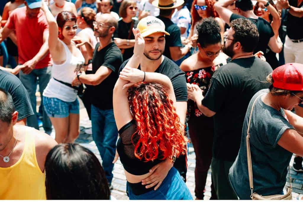 A woman with curly red long hair in a black tee shirt and jeans dancing with her arms up, with a man with a white baseball cap and a beard.