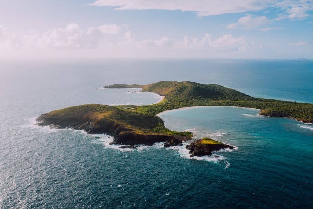 An aerial view of a beautiful green island with waves crashing on its shores, against a deep turquoise sea.