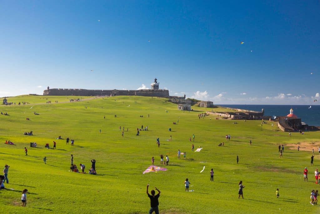 A large green lawn with an ancient building in the background and the ocean to the right.  The people on the lawn are hanging out and some are flying kites.