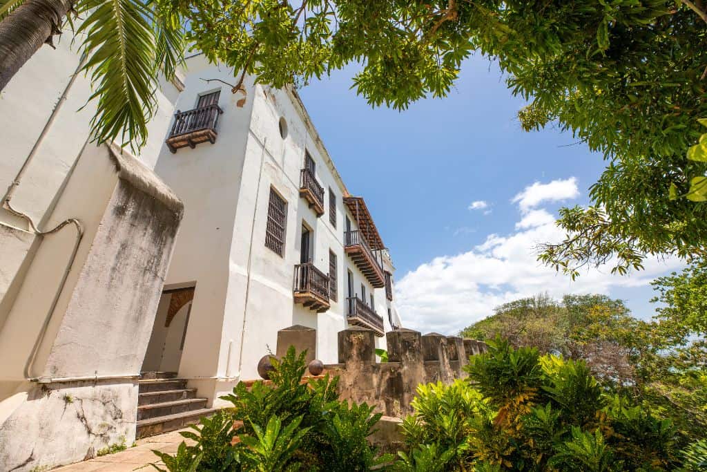 An old white building with  some windows and balconies and weathering on the side of it, surrounded by an old looking stone fence and brightly green colored tropical trees.