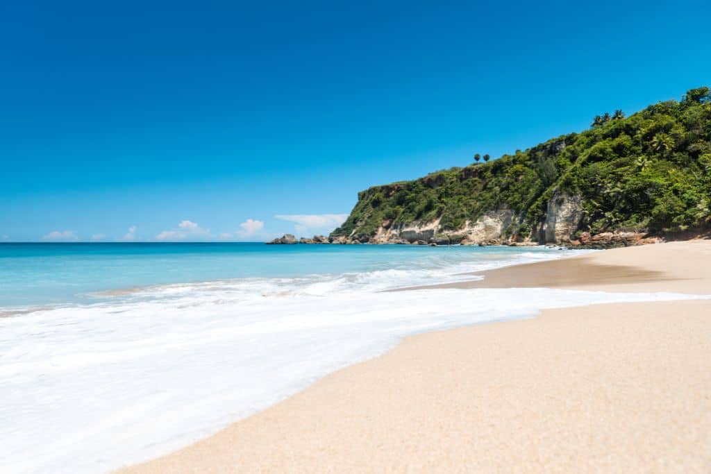 A white sand beach with calm turquoise water and a cliff with tropical plants in the distance.