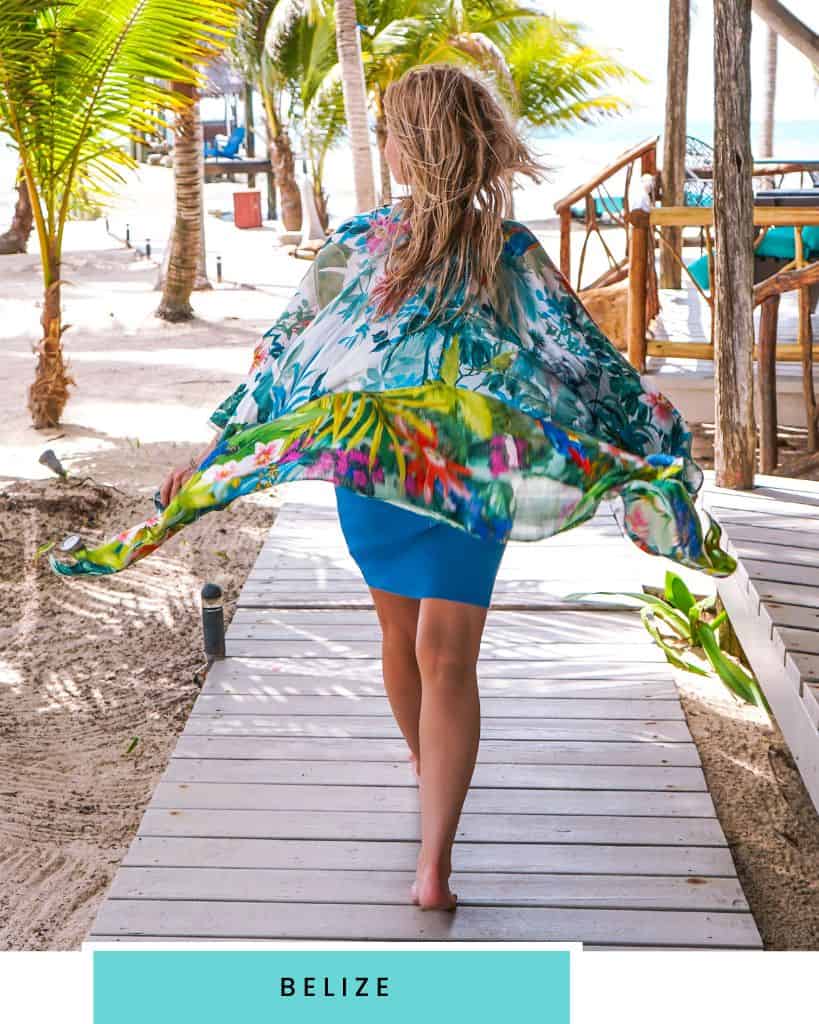 blonde woman wearing a brightly colored flower kimono and blue dress walking down a boardwalk with palm trees around her