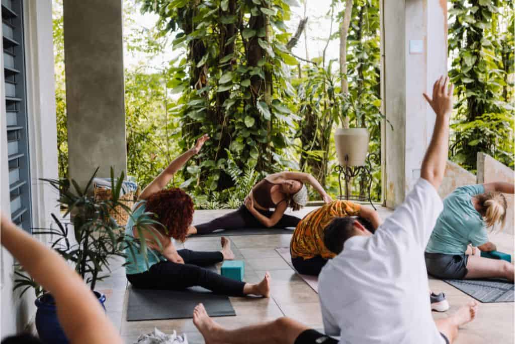 6 people doing seated side body stretches on an outdoor deck in a yoga class with a jungle behind them.