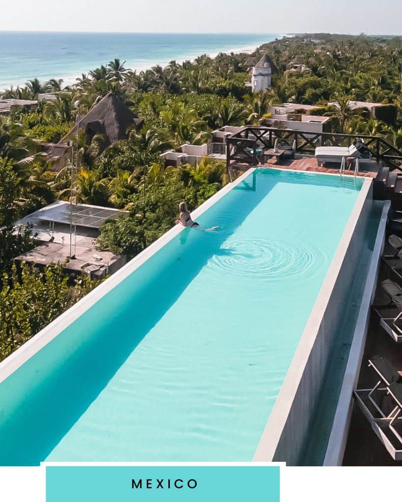 A large rectangular brightly blue colored pool with a blonde woman looking out over the edge surrounded by tropical trees and the ocean