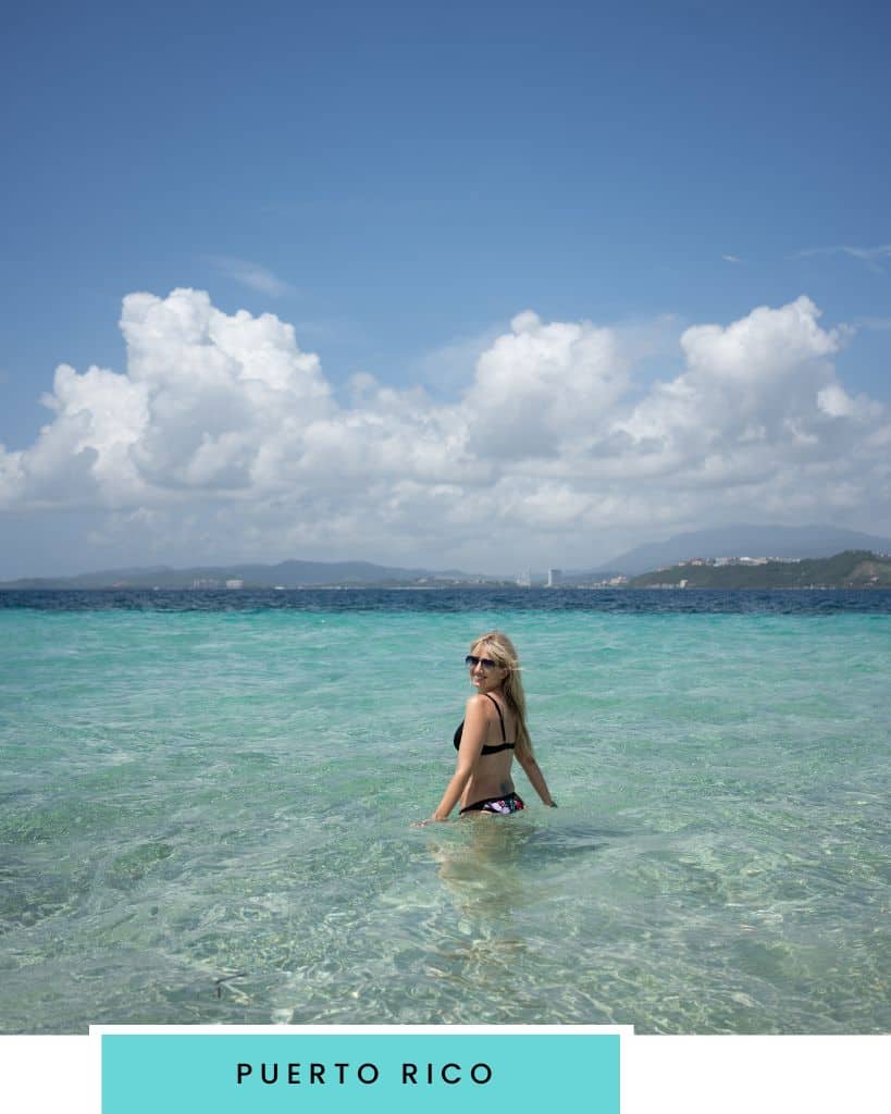 blonde girl in bikini standing in a calm clear turquoise ocean looking over her shoulder towards the camera with mountains and a city in the background