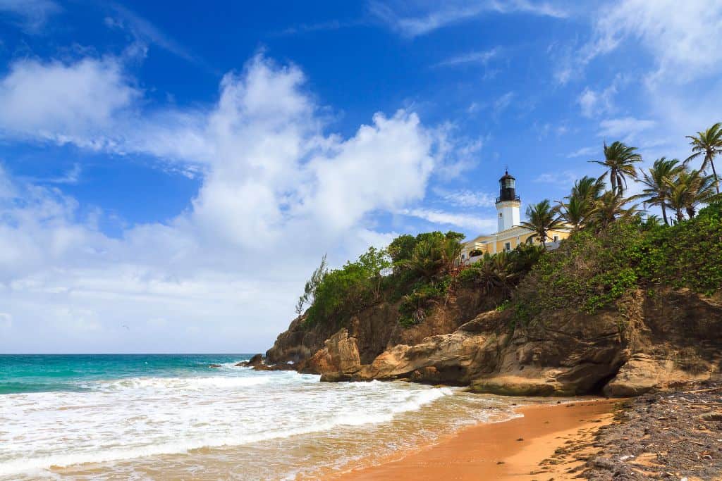 Tall cliffs with trees on them plunging down into a turquoise ocean, with a white, yellow and black lighthouse sitting on top of the cliff.