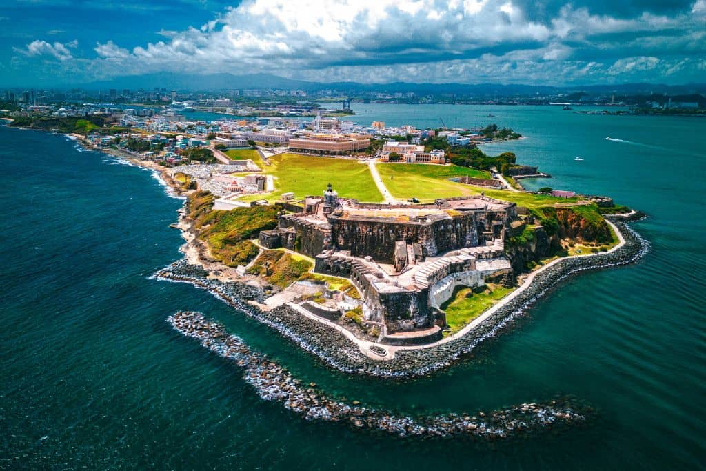 A brightly colored aerial view of a historic fort sitting in a bright green lawn on an island with a city behind it.