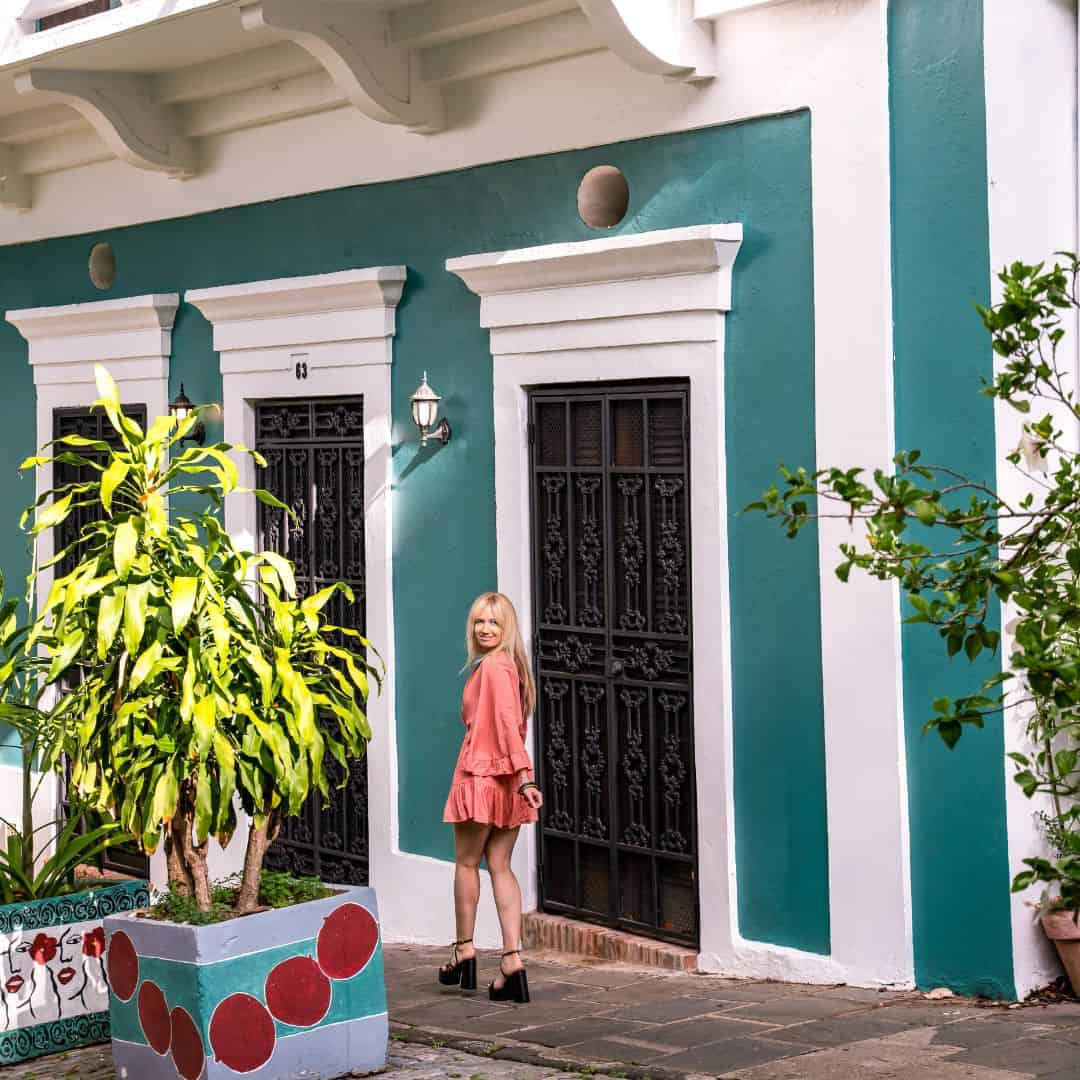 Woman with long blonde hair wearing a peach shorts jumpsuit and black heels looking over her shoulder and smiling as she's walking down a street in front of a dark turquoise building with three black doors lined with white frames and tropical trees in brightly painted pots.