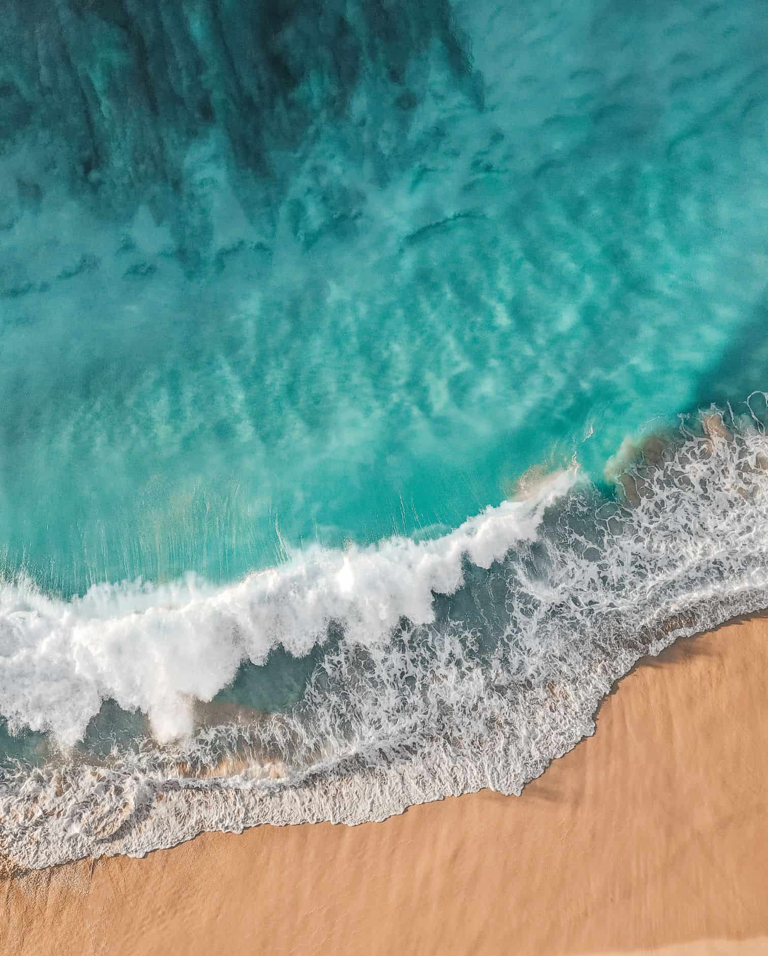 An aerial shot of a brightly colored turquoise ocean and white waves crashing along a warmly colored sandy shore.