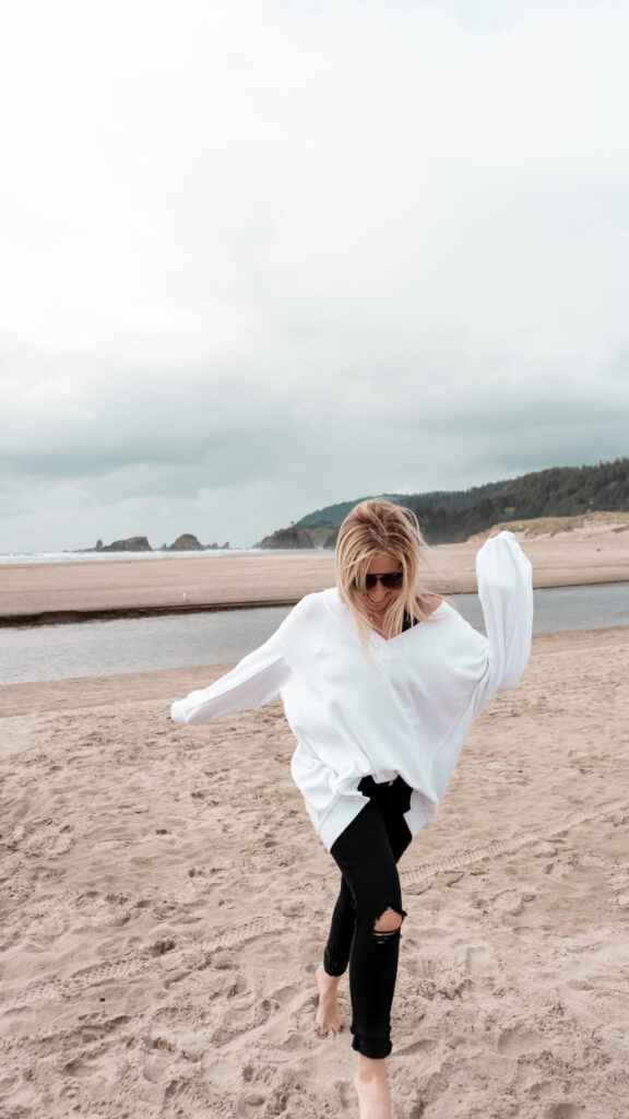 Blonde woman wearing black sunglasses, a white sweatshirt and black jeans running towards the camera with a moody ocean behind her.