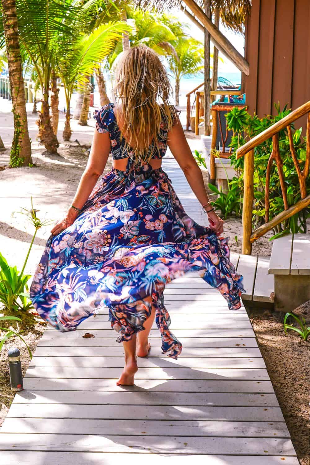 Blonde woman wearing a long bloe dress with brightly colored flowers on it walking down a wooden sidewalk with sand and palm trees on the left and staircases up to hotel rooms on the right.