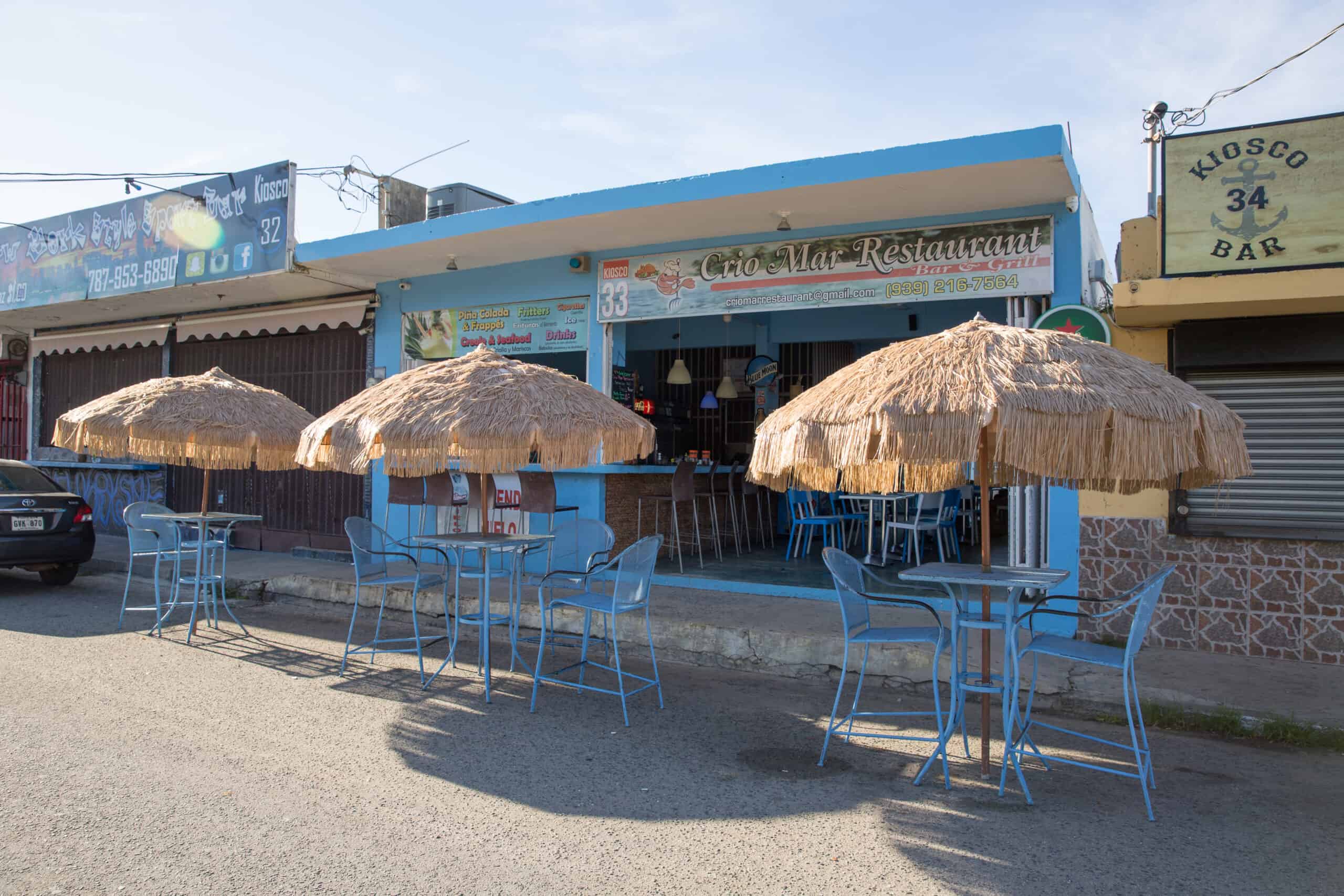 A strip of several restaurants and bars with signs on them, lined with streetside tables and tiki umbrellas. 