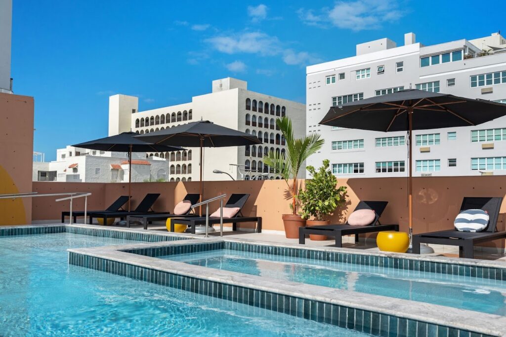 A brightly colored blue pool and hot tub lined with black lounge chairs, umbrellas and tropical plants overlooking a city with white buildings under a bright blue sky.