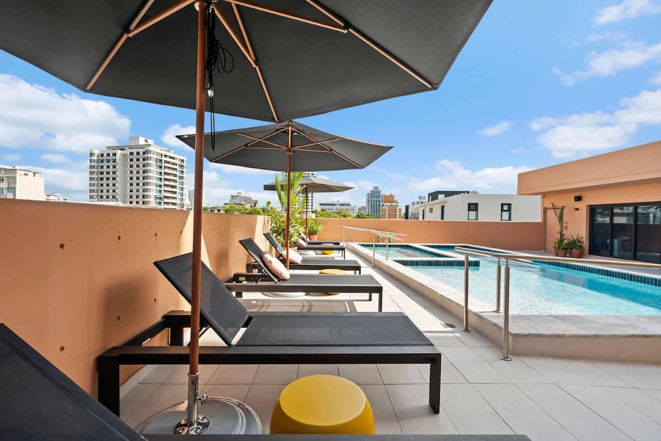 A line of black lounge chairs with black umbrellas over them lining a pool deck surrounded by orange walls.