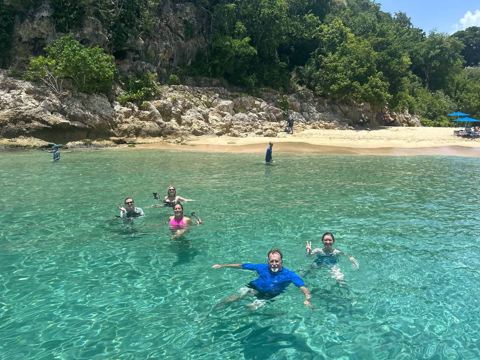 A group of 5 swimmers sitting in clear turquoise ocean water just off the shore, waving hi to the camera