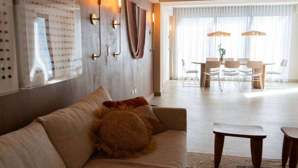 A view looking into a dining room near a bright window over a couch with red toned pillows and light fixtures on the wall