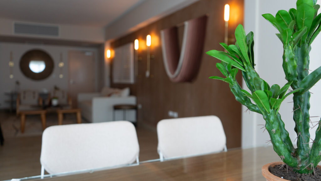 A view from a table with a plant on it and white chairs at it of a hotel room with a pink wall tapestry and lights on the wall