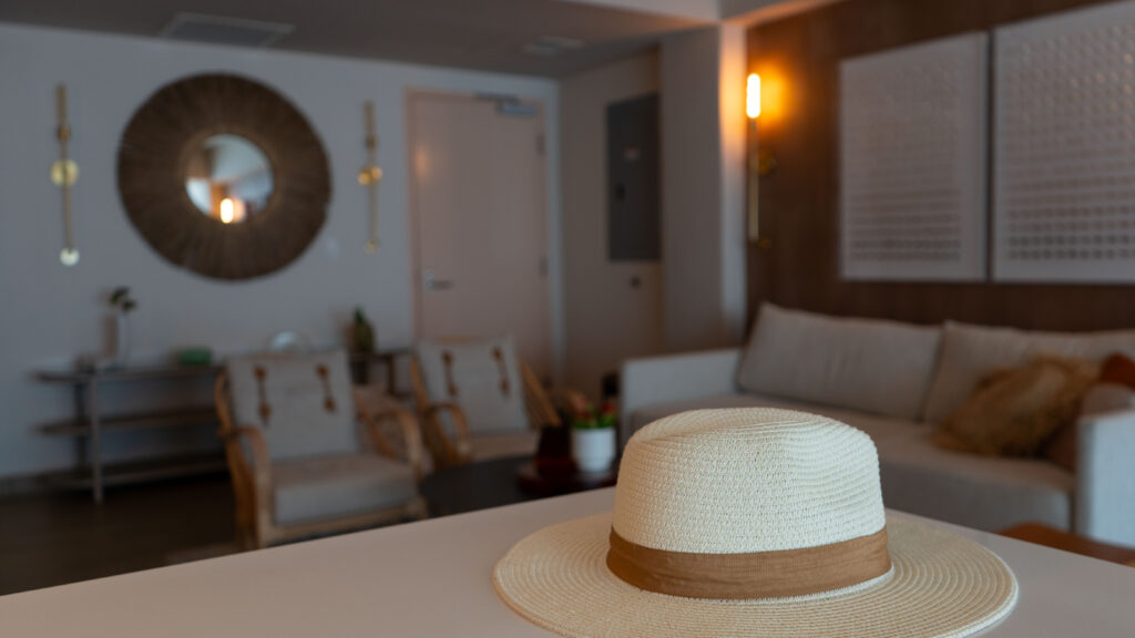 A white beach hat with brown stripe in the foreground with a living room with two white chairs and a couch and a pink door in the background.