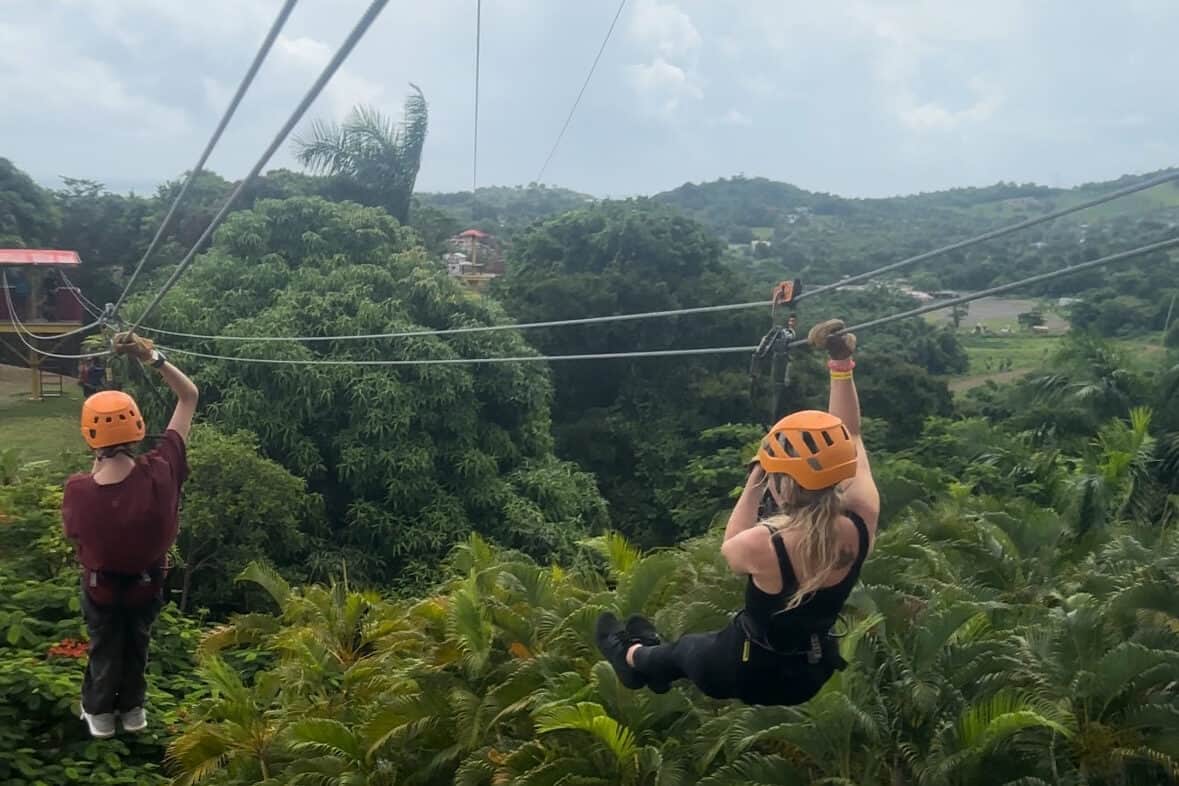 A child in a red tee shirt and a woman with blonde hair wearing black, ziplining through a rainforest and wearing orange helmets.