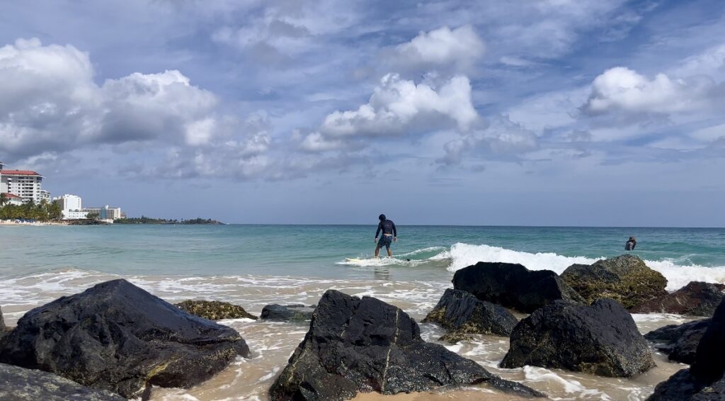 A man wearing a dark shirt surfing on a white board along a rocky shore