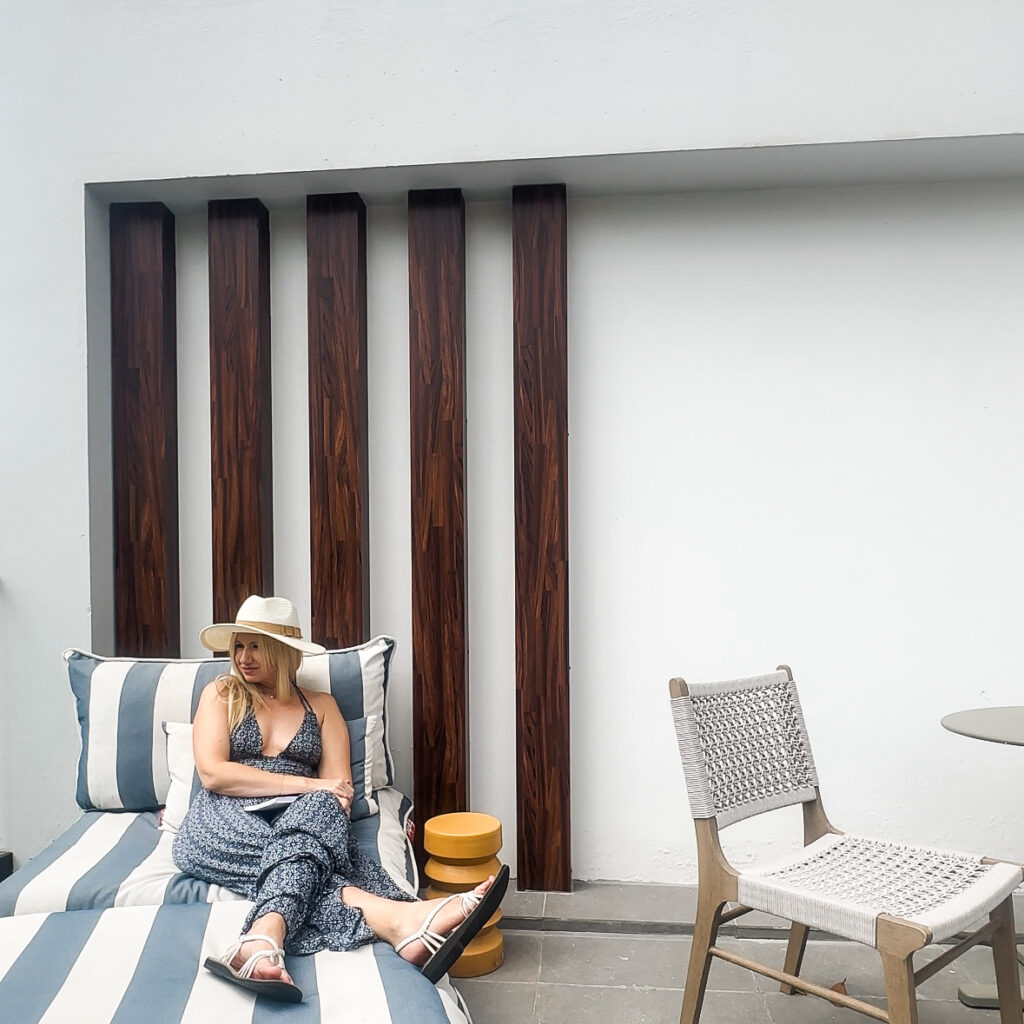 A blonde woman in a long blue and white dress wearing a white beach hat sitting on a blue and white lounge chair with a white table and chair next to her.