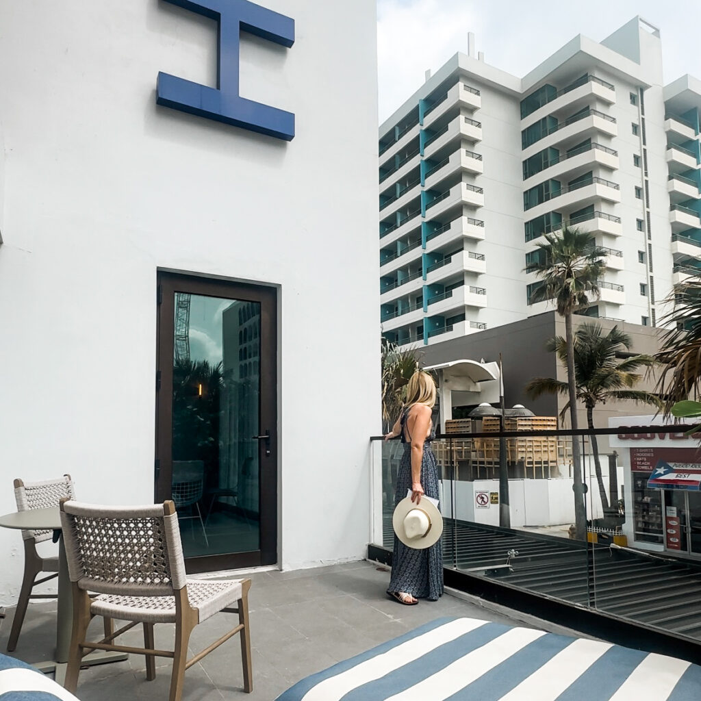 A blonde woman in a long blue and white dress holding a white beach looking out at a tropical cityscape form a balcony.