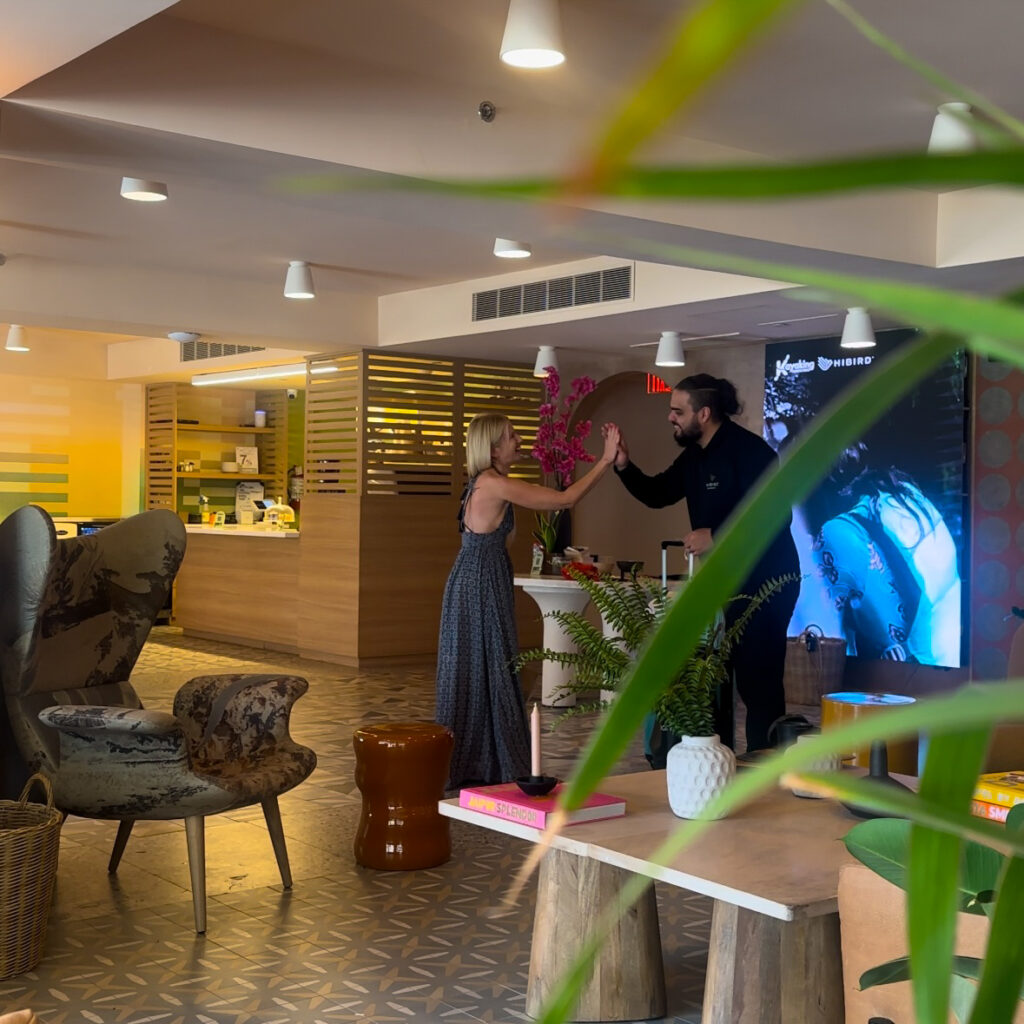 A blonde woman in a blue dress high-fiving a man in all black in a hotel lobby.