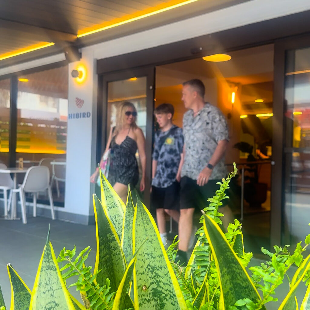 A blonde woman, boy and man in a white and black shirt walking out of a building called Hibird with snake plants in the foreground.