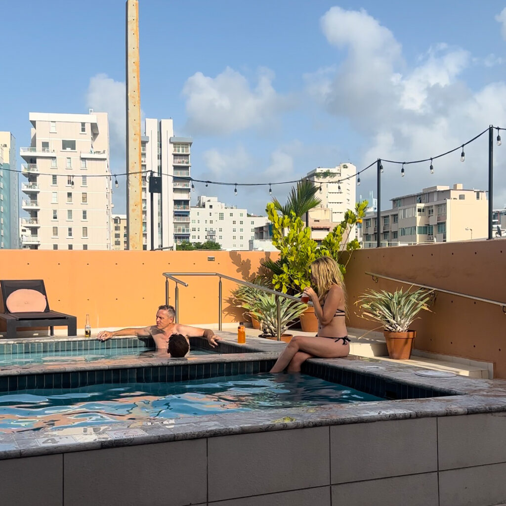 A woman sitting on the side of a pool sipping on a drink with a man and boy in the pool, plants and a city behind them