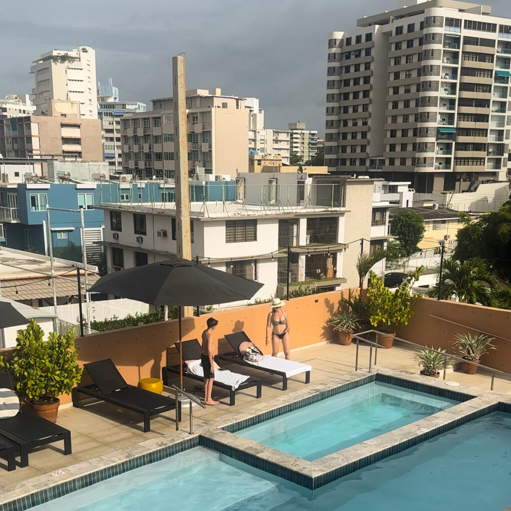 An aerial shot overlooking a pool with umbrellas and chairs on the deck and a woman and boy putting their towels down and a city behind them.