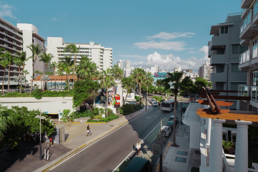 An overhead shot of a city with a street running down the center, palm trees lining the street and people walking on the sidewalk.