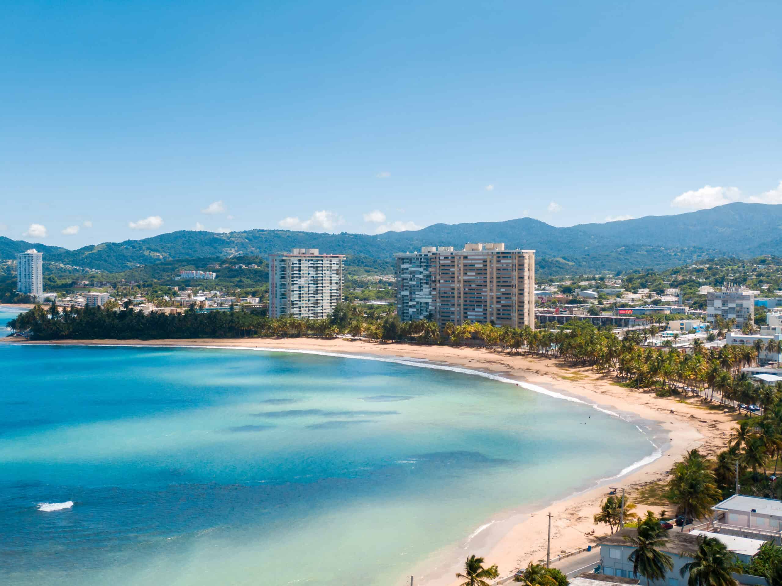 A turquoise ocean bay lined with sand, palm trees and a small city with some highrises and a busy town