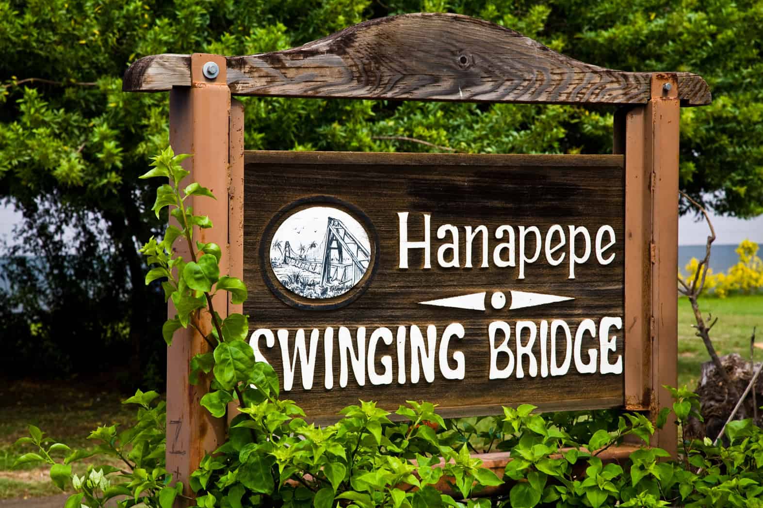 A wooden sign surrounded by trees for Hanapepe Swinging Bridge