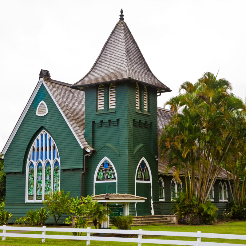 A tall green church with beautiful stained glass windows and a white picket fence surrounding it in Hanalei