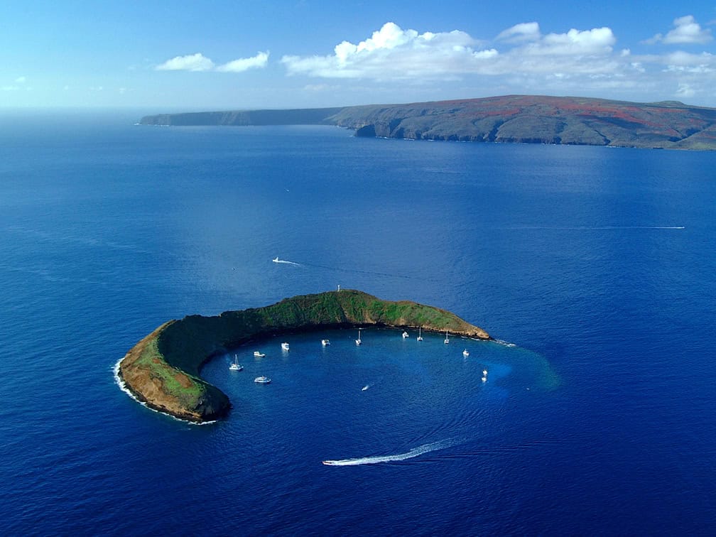 A stunning aerial shot of a crescent shaped island in a deep blue ocean, sailboats around it and another island in the background