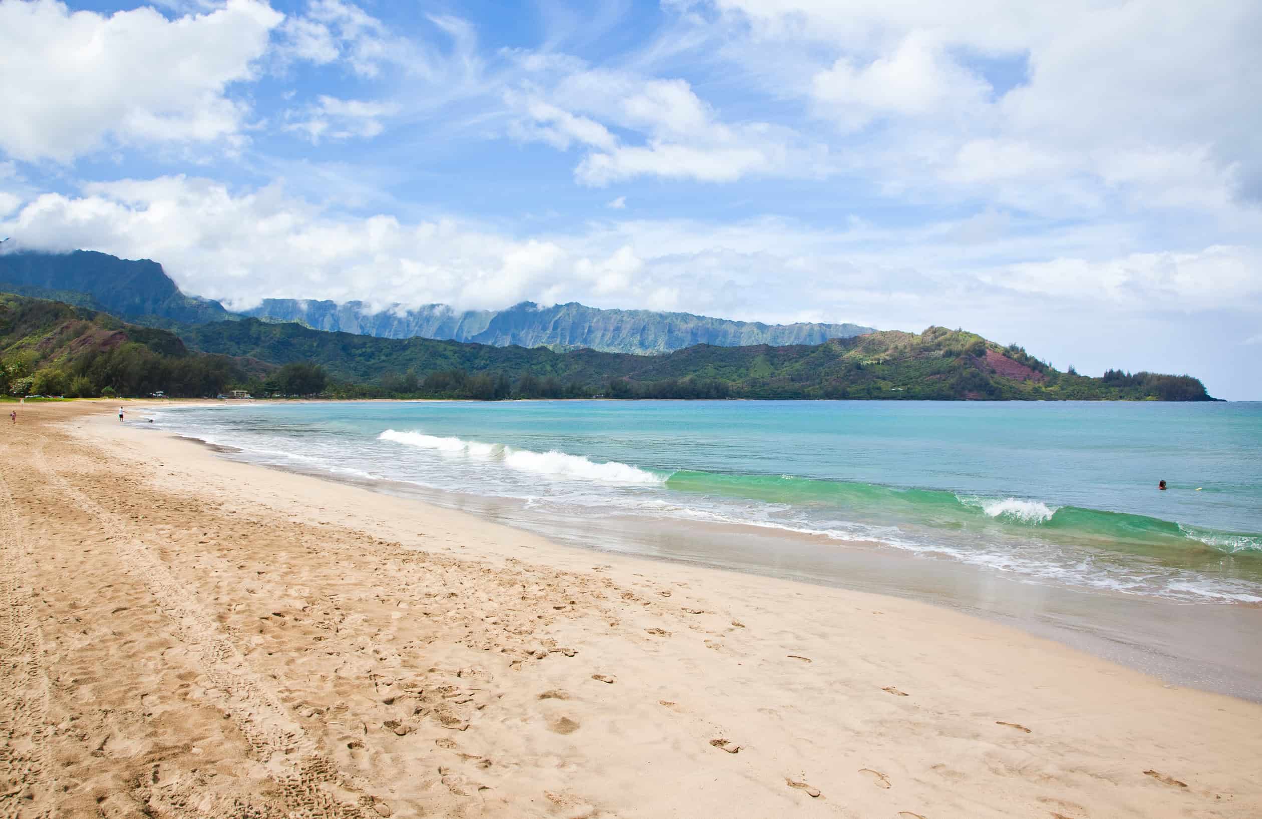 A white sand beach with calm waves and a mountainous landscape in the background