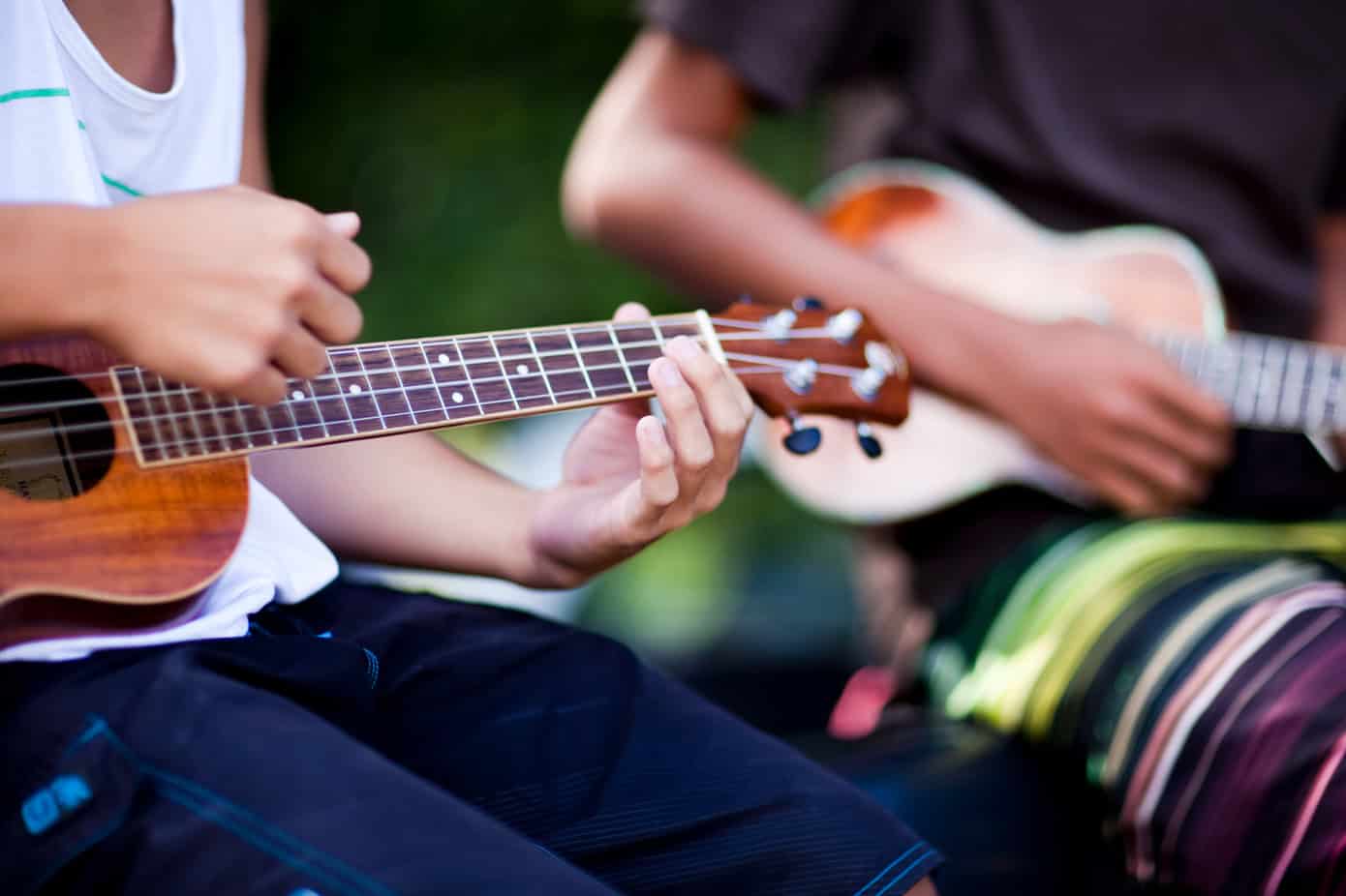 An up close shot of a woman in a white tee shirt and navy blue pants and a man in a brown tee shirt and colorful shorts playing the ukulele