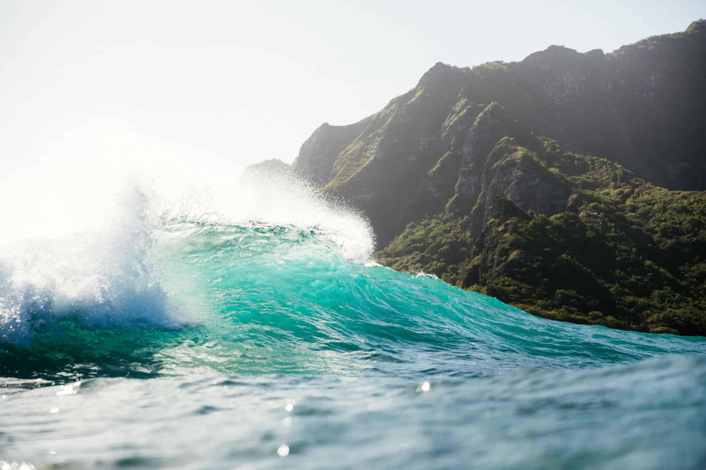 A wave of clear turquoise water crashing with a green mountain in the background.