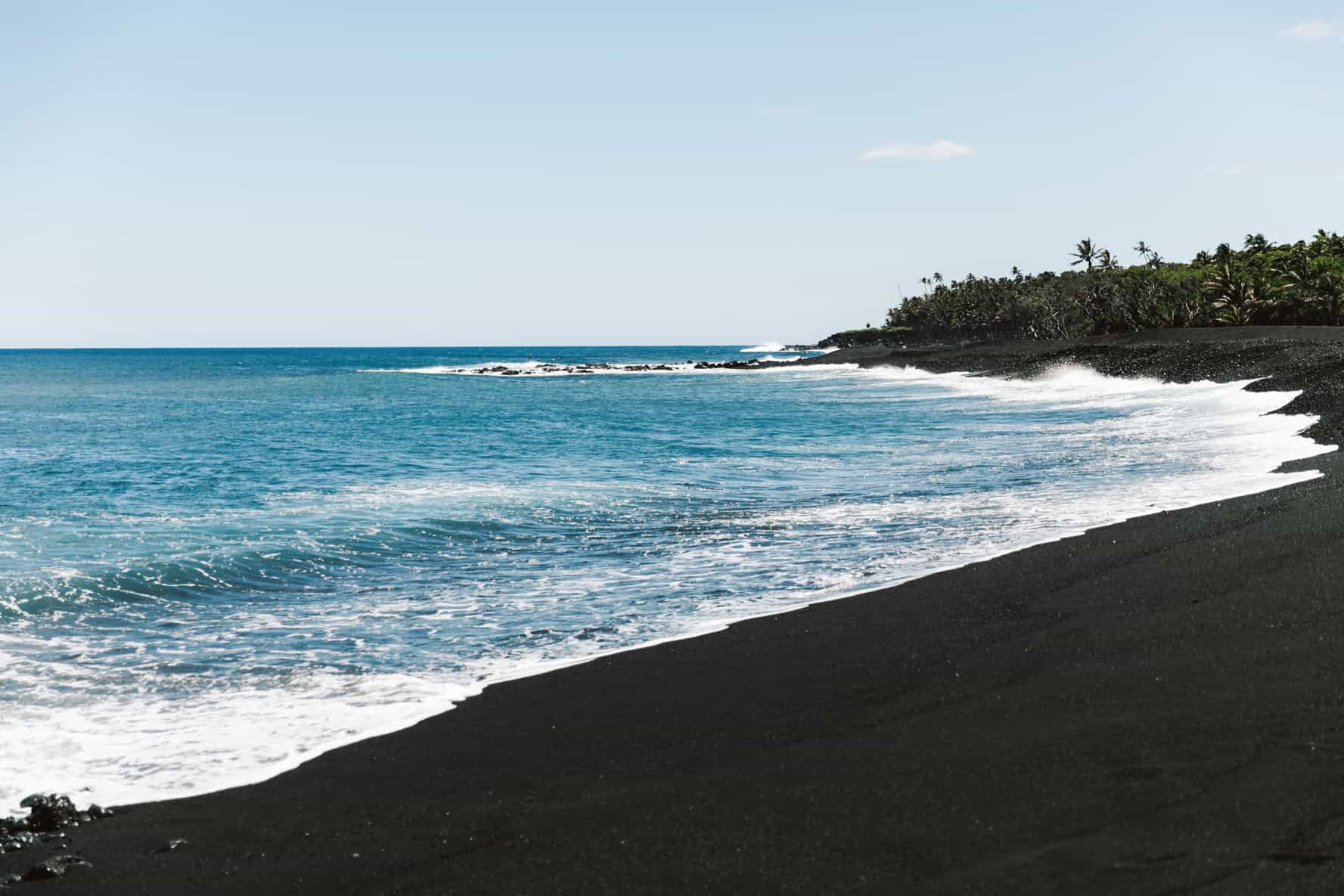 A calm turquoise ocean agains a black sand beach with trees in the background