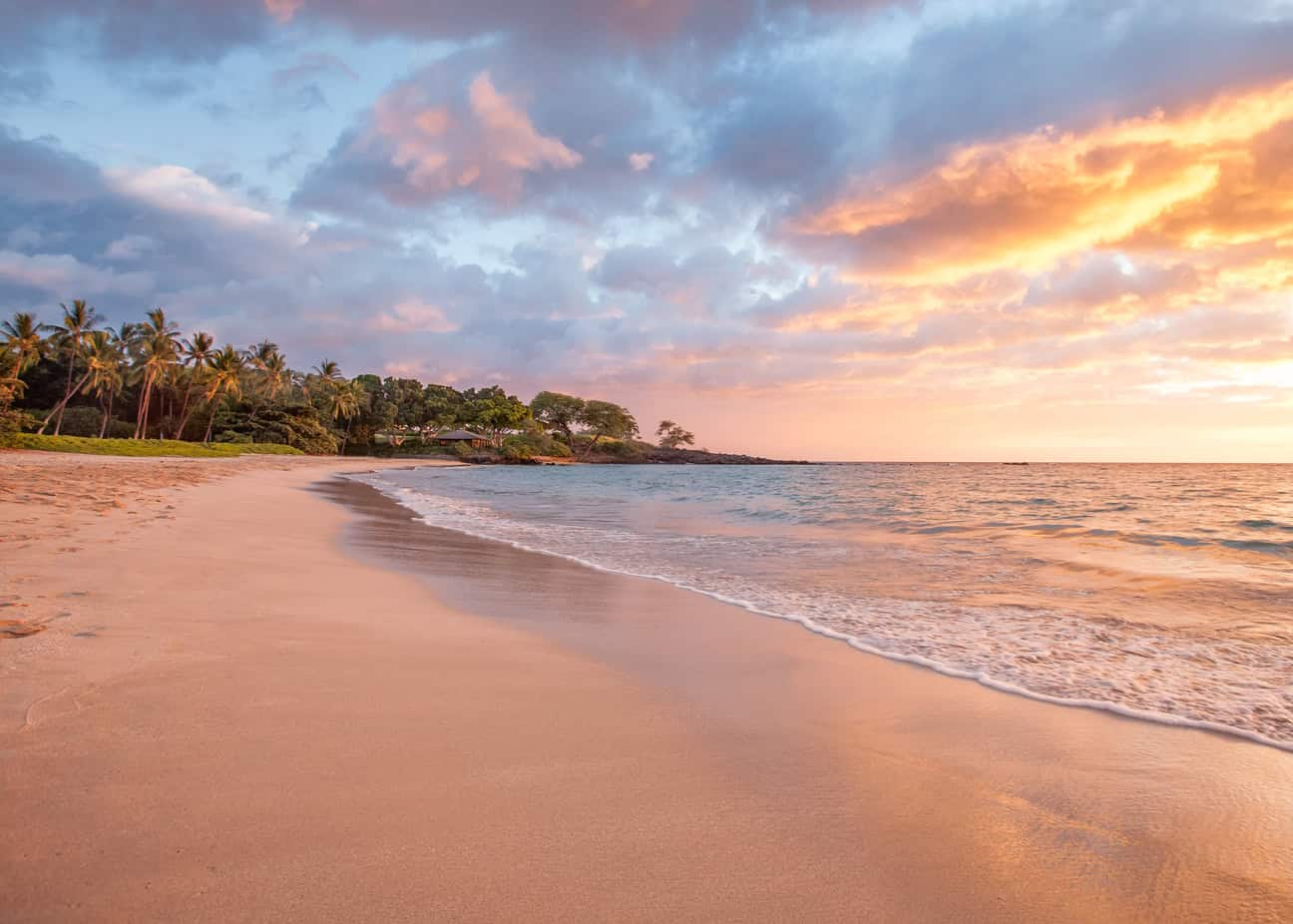 A sandy beach with calm waters and trees to the left with a brightly colored sunset