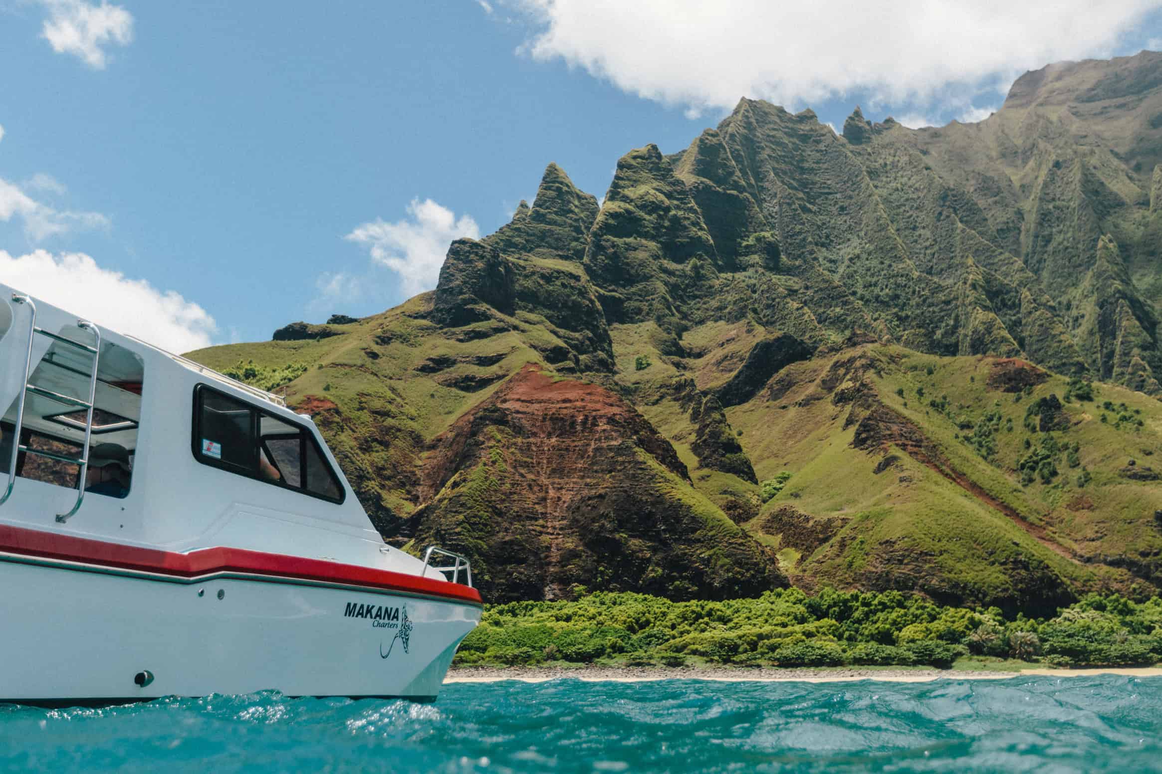 A white boat with a red stripe sitting in turquoise ocean water, facing a tall mountainous island with tropical plants on it