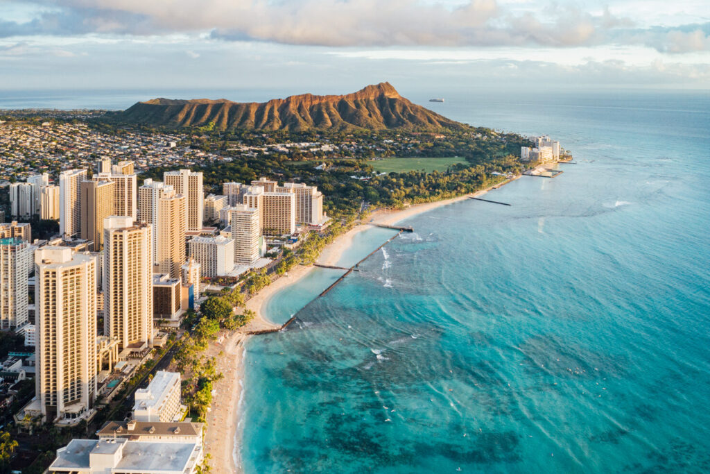 An aerial view of Honolulu with the city in the foreground, Waikiki beach, clear ocean waters and mountains in the background