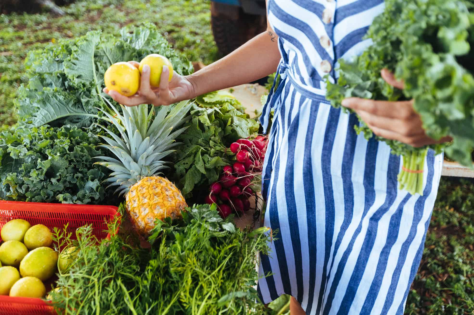 A women in a blue and white striped outfit picking up fruits and veggies in a farmers market in Kauai