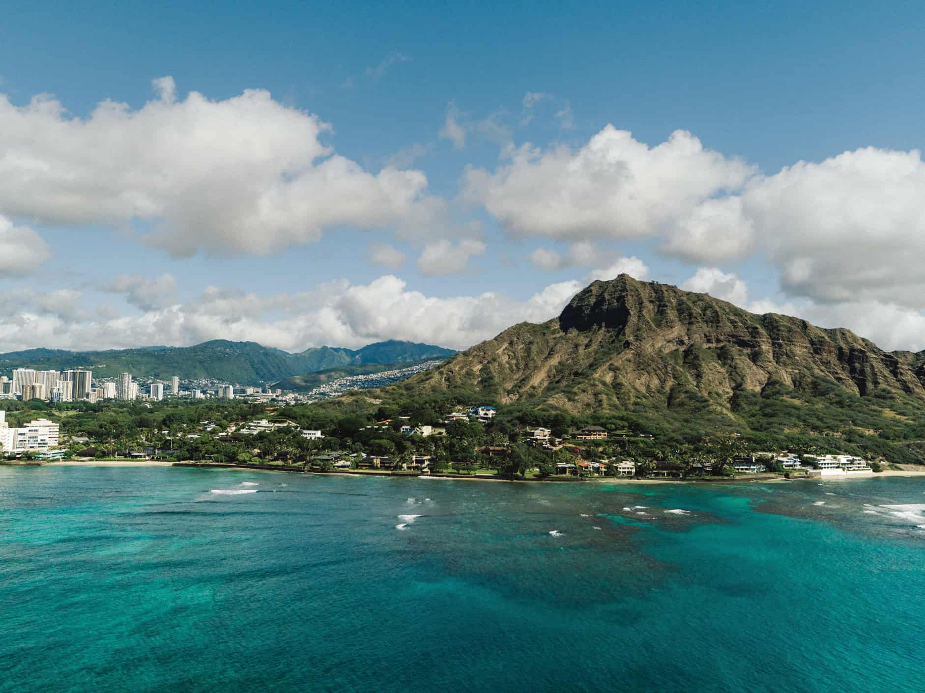 A shot over the bright turquoise ocean looking back towards land with a mountain in the foreground and house in front of it, and a city and more mountains in the background.