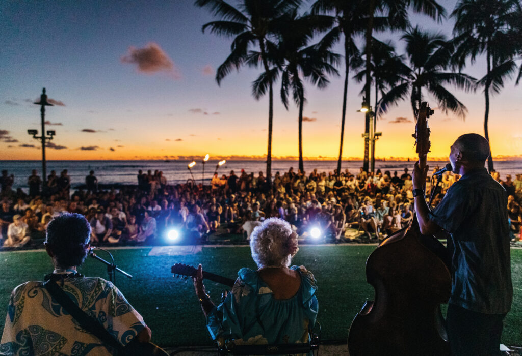 The backs of three musicians holding guitars looking out to a crowd of people with the ocean and palm trees behind them at sunset